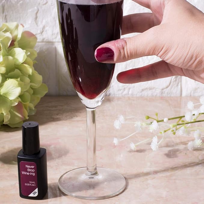 Mano con uñas burdeos cogiendo copa de vino junto a frasco de esmalte de gel para uñas Never Stop Wine-ing