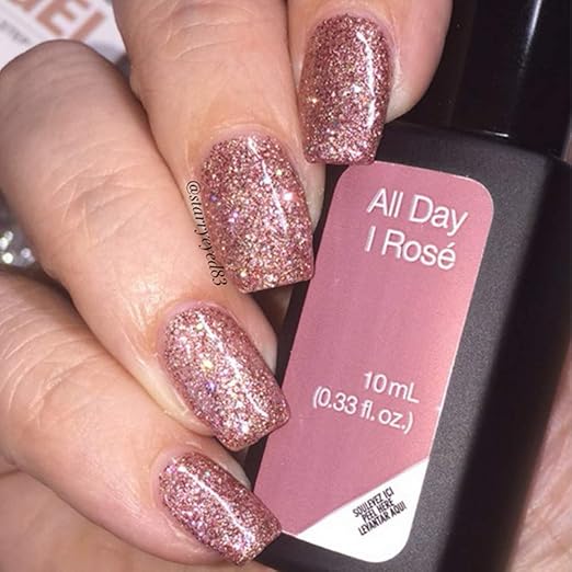 Mano femenina con uñas rosas con brillantina sosteniendo esmalte de gel para uñas semipermanentes color rosa All Day I Rose